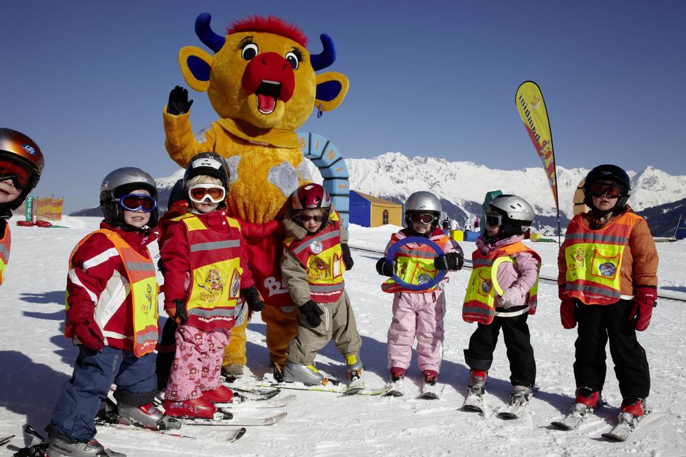 Auf dem Bild ist eine Gruppe von Kindern in Skikleidung zu sehen. Hinter ihnen steht das Maskottchen "Berta" (eine verkleidete Kuh).